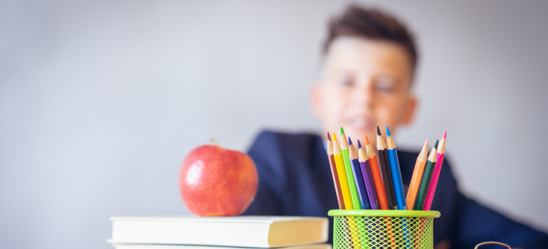 A boy that is in the background out of focus is looking at a table with books, pencils, and an apple on it and smiling as he is happy that he is moving to Orange from Newport Beach.