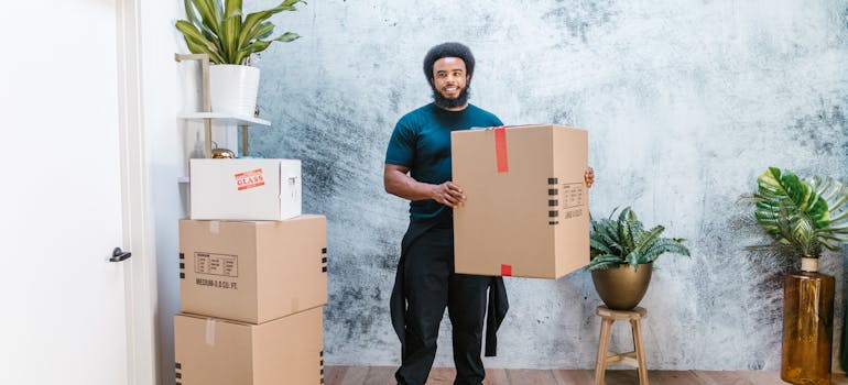 A man working as a professional mover who is wearing a green shirt is holding a cardboard box.