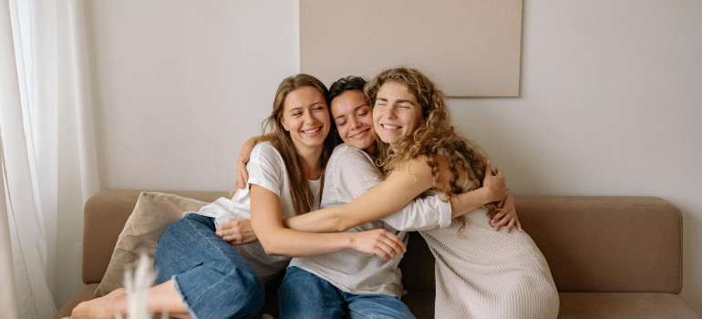 Three friends sitting on a couch, smiling and hugging each other.