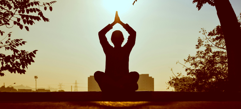 Person meditating at sunset with a cityscape backdrop. A self-care tip to build a new routine after moving.