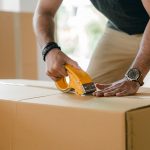 a man sealing a cardboard box with a duct tape because he is moving for a job