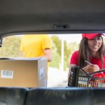 Woman loading groceries and a box into the back of a car while checking a list.