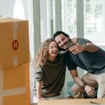 A smiling couple taking a selfie surrounded by moving boxes in their new home.